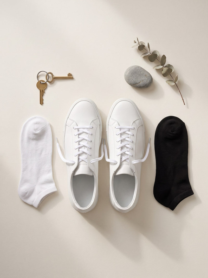 A white and a black Trainer Sock laid flat either side of a pair of white leather trainers