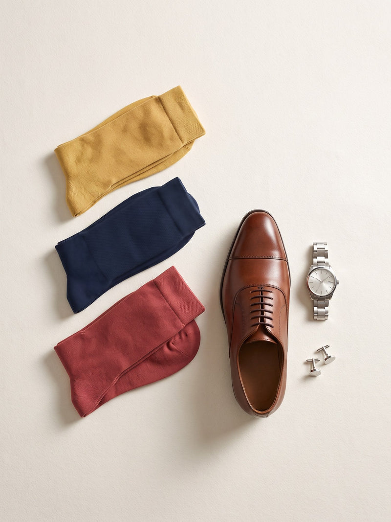 Mustard, navy and brick Silver Edition dress socks laid flat beside a brown oxford shoe and a watch
