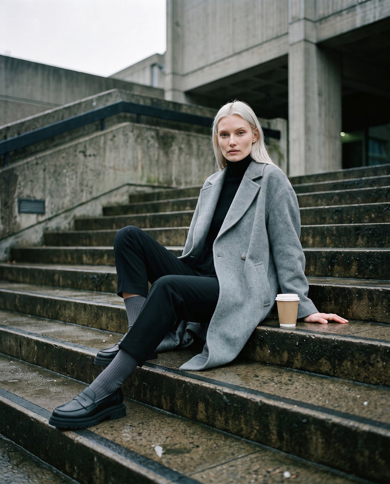 A woman in a grey coat on stone steps in London, charcoal socks and loafers