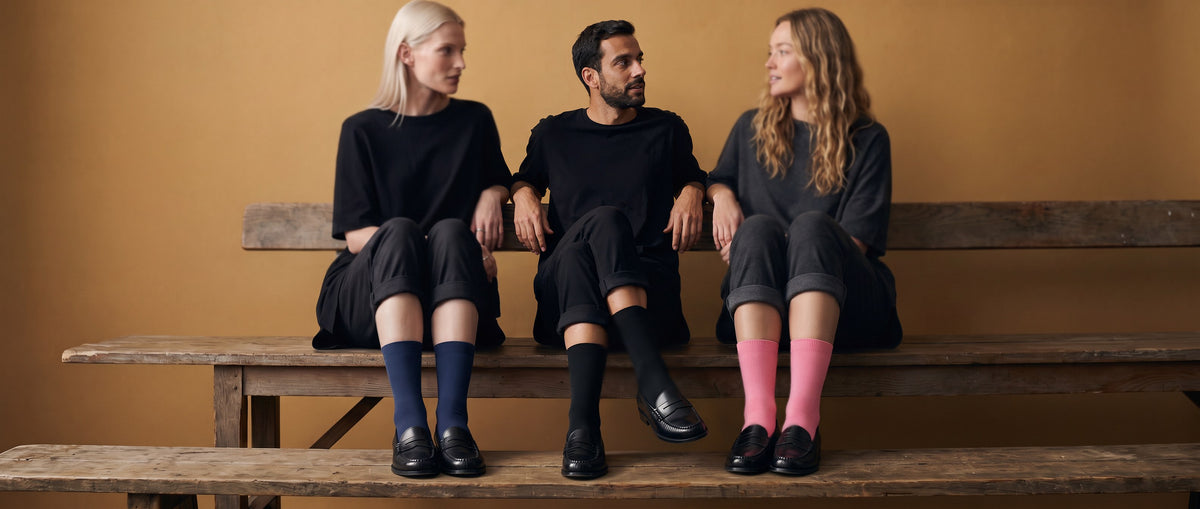 Two women and a man in black sit on a wooden bench against an ochre wall, in navy, black and pink Silver Edition socks with black penny loafers