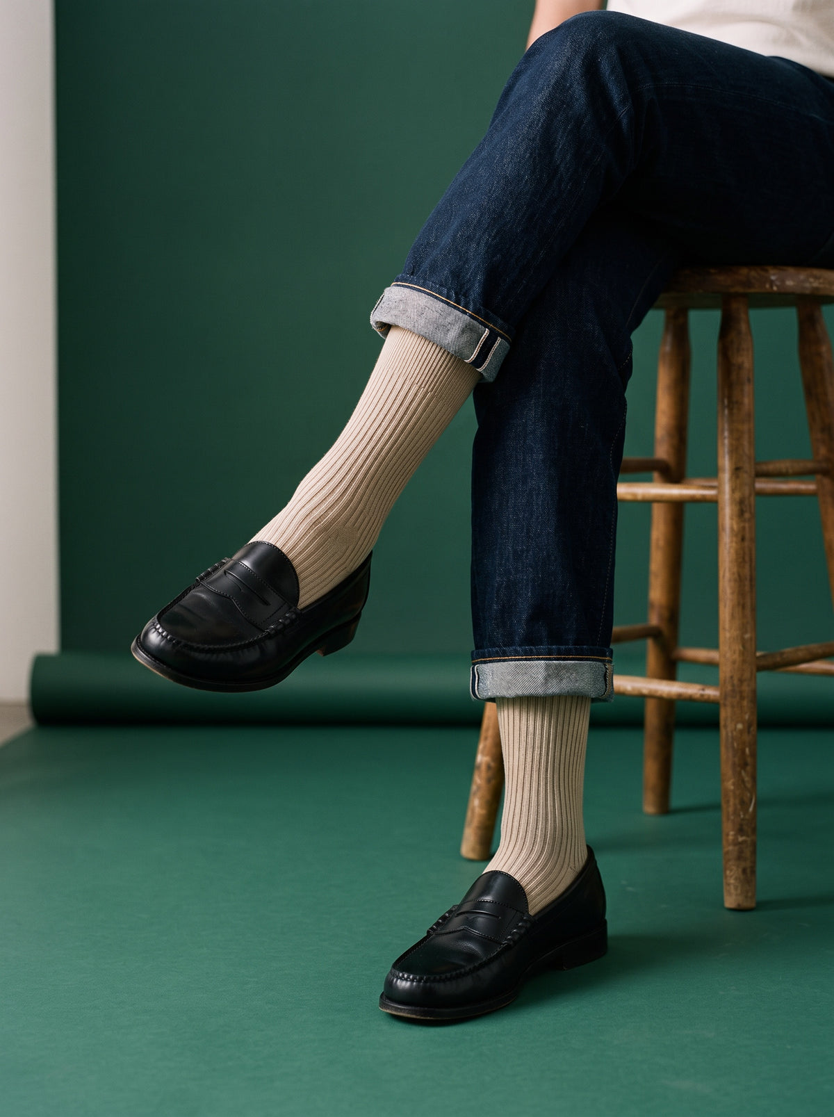 Sand ribbed crew socks worn with black loafers and turned-up jeans, seated on a stool