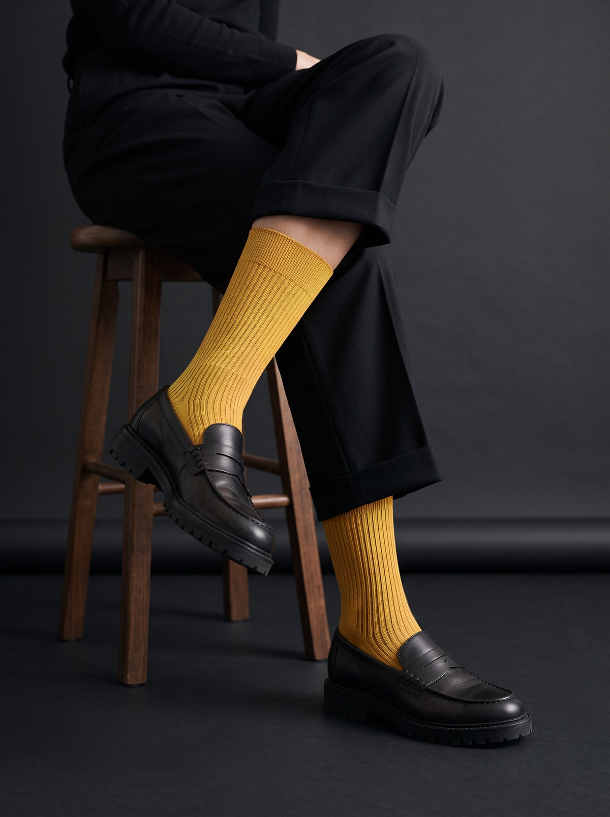 Ochre ribbed crew socks worn with black loafers and black trousers, seated on a stool