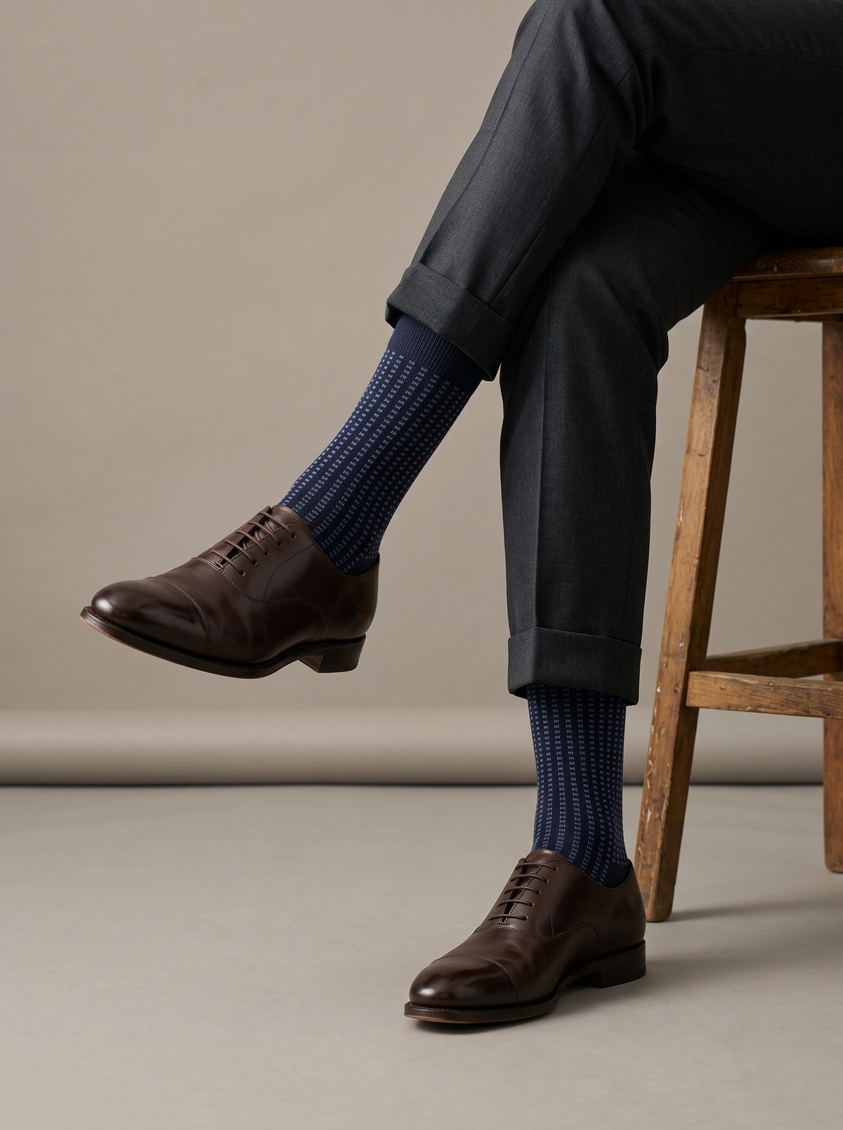 Navy jacquard crew socks worn with brown brogues and charcoal trousers, seated on a stool